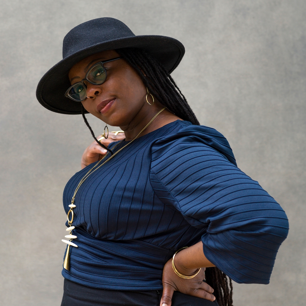 Black woman wears blue long sleeve top and black hat and glasses and poses facing left with hand on hip and head tilted toward camera